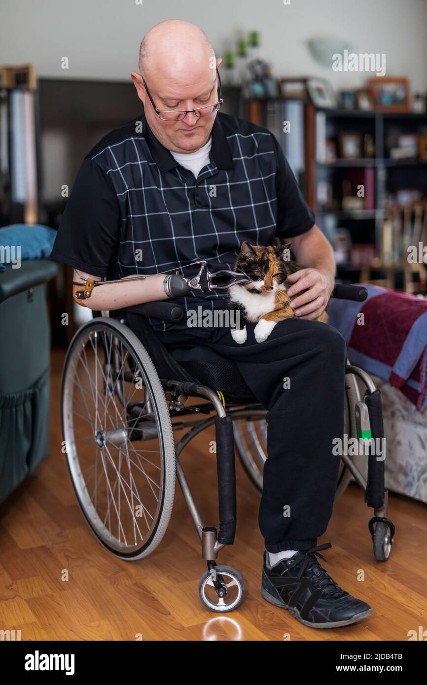 Ein Mann mit einer Amputation von zwei Gliedmaßen sitzt in seinem Haus mit seinem Haustier auf dem Schoß Albert, Alberta, Kanada Stockfoto