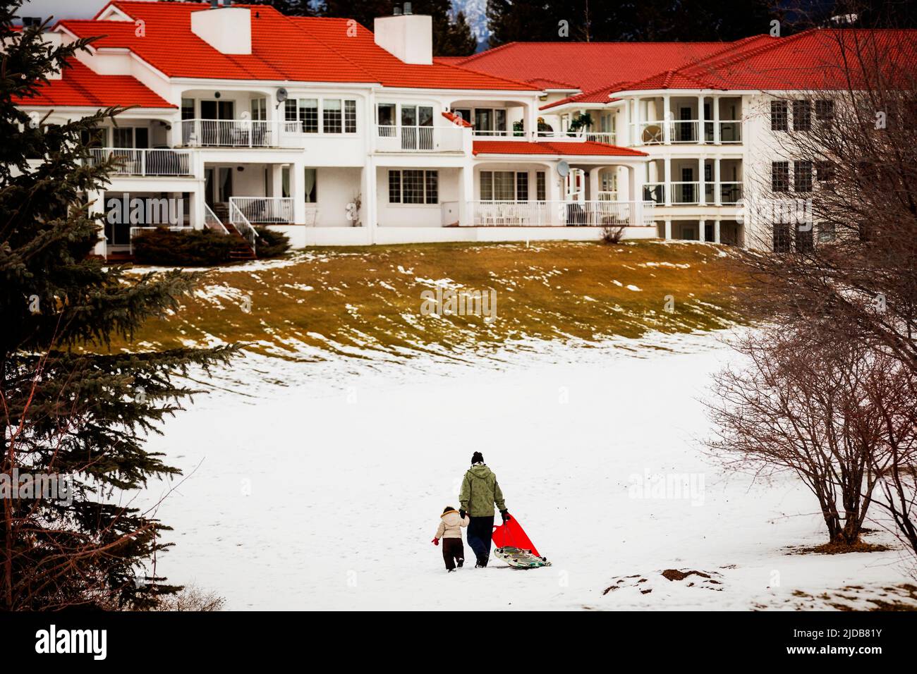 Eine Mutter hält die Hand ihrer jungen Tochter, während sie Schlitten auf Einen Snowy Hill ziehen und zu Einem Gebäude in Einem Mountain Resort gehen; Fairmont, BC, Kanada Stockfoto