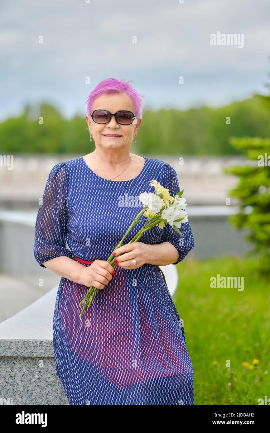 Porträt einer lächelnden reifen Frau mit kurzen rosafarbenen Haaren, die in der Nähe von Tannenbäumen auf der Straße steht Stockfoto