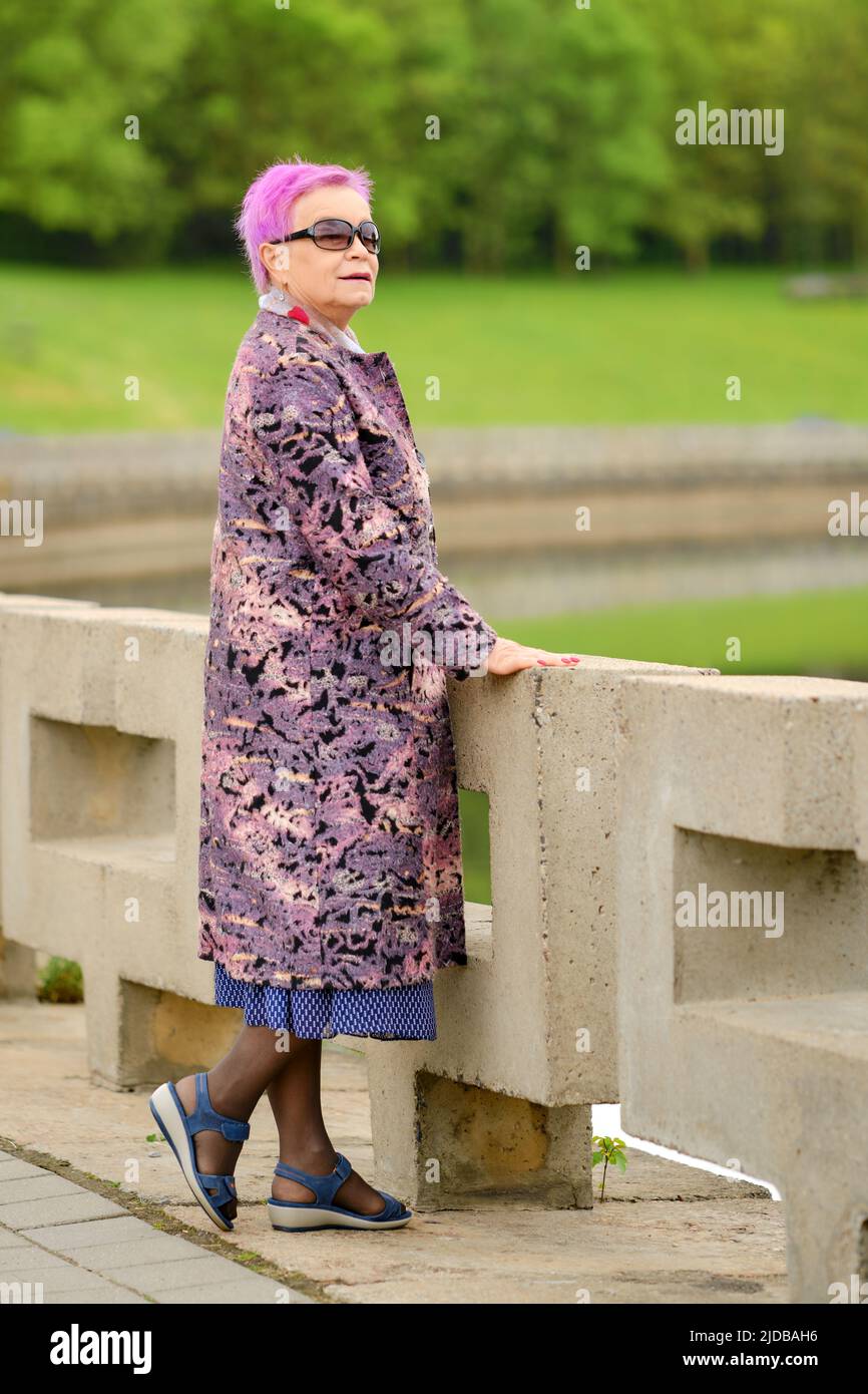 Porträt einer alten Frau mit kurzen rosafarbenen Haaren und Wollmantel, die am Flussufer im Stadtpark steht Stockfoto