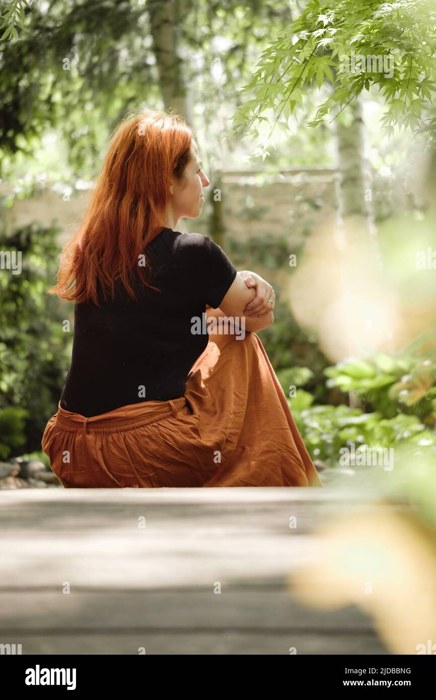Frau mit 40s Jahren sitzt auf dem Boden und blickt weg in einen grünen Garten. Ruhe haben, denken. Weibchen mit Ingwerhaar von hinten auf der Brücke Stockfoto