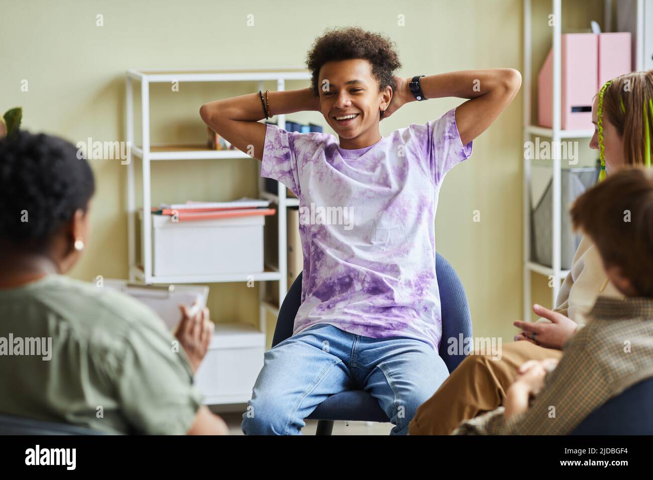 Afrikanischer Teenager, der auf einem Stuhl sitzt und lächelt und sich während einer psychologischen Sitzung mit der Gruppe und dem Psychologen schlecht benommen hat Stockfoto