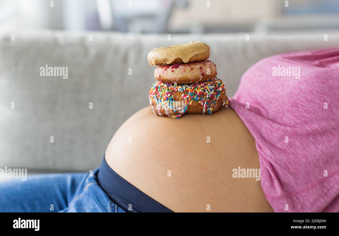 Schwanger Frau mit Donuts auf dem Bauch. Heißhunger auf Desserts und Süßigkeiten während der Schwangerschaft, Mädchen essen ungesunde Backwaren auf Baby Bump für Gestationszeit Stockfoto