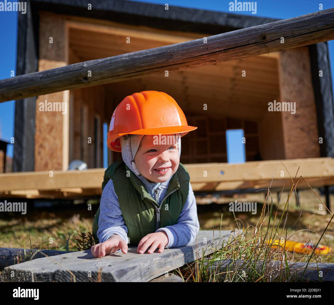 Porträt eines Kleinkindes, das am sonnigen Tag als Baumeister auf der Baustelle spielt. Kinderschreinerin in orangefarbenem Helm lächelt auf Holzrahmen im Hintergrund des Hauses. Schreinerei-Konzept. Stockfoto