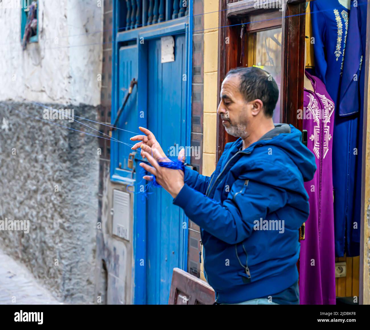 Ein Mann, der an Strick-Fäden arbeitet - Strickmaterial für die professionelle Bekleidungsherstellung vor seinem Bekleidungsgeschäft. Essaouira. Marokko Stockfoto