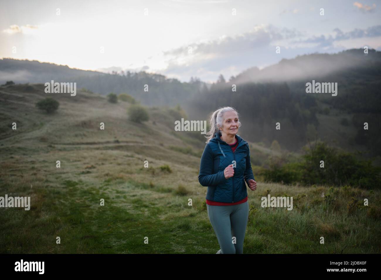 Ältere Frau joggt am frühen Morgen in der Natur mit Nebel und Bergen im Hintergrund. Stockfoto