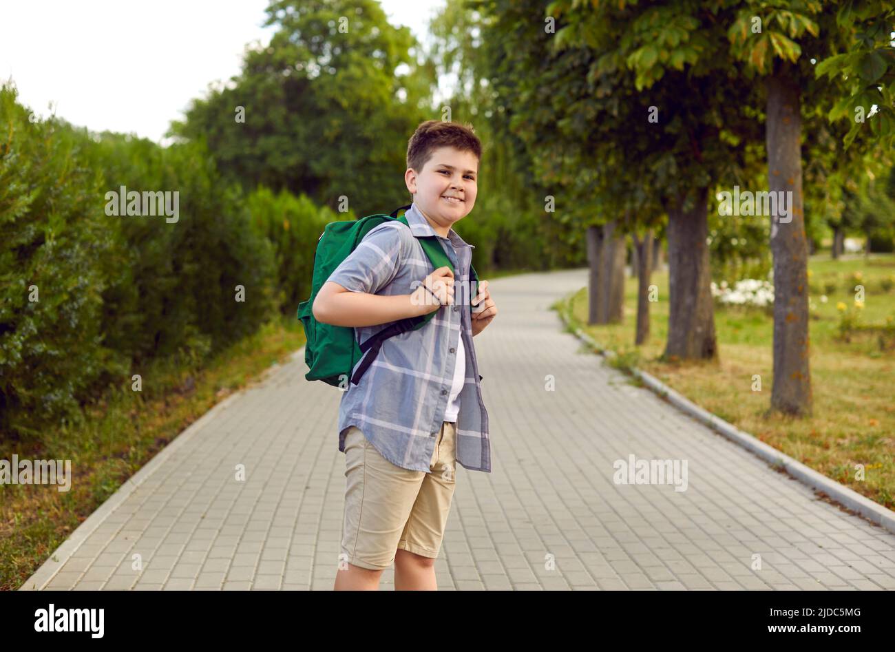 Porträt eines lächelnden Jungen, der am warmen Morgen mit seinem Schulrucksack in den Park zur Schule geht. Stockfoto