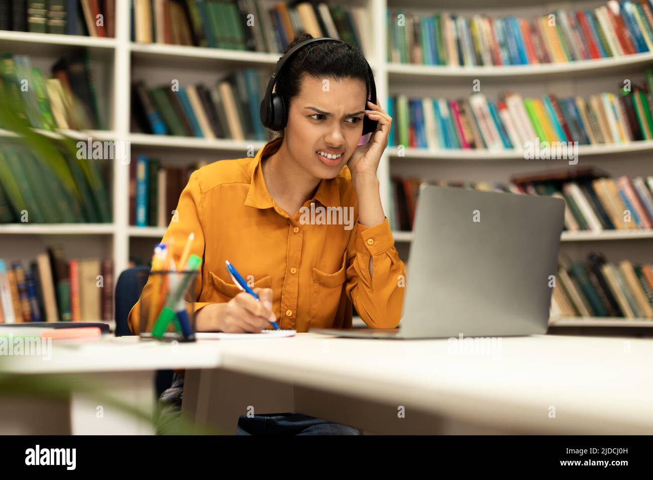 Die junge Frau, die mit einem Laptop online studiert und Hörprobleme in ihren Kopfhörern hat, sitzt im Inneren der Bibliothek Stockfoto