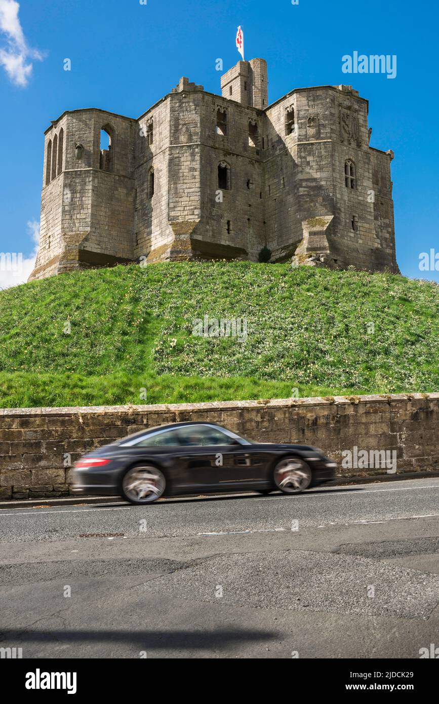 Autofahren, Blick im Sommer auf einen Sportwagen, der an einer verfallenen mittelalterlichen Burg in der englischen Landschaft vorbeirast, Warkworth, Northumberland, Großbritannien Stockfoto