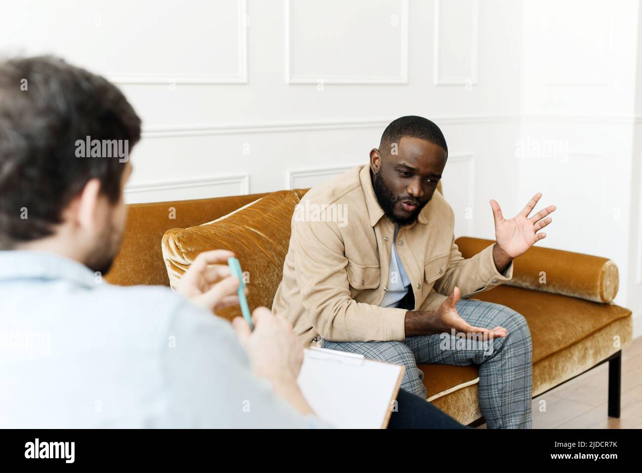 Ein irritiert Afroamerikaner spricht emotional mit einem Psychologen über Probleme bei einer Sitzung, ein verwirrter unglücklicher junger Mann wird mit einem therapiert Stockfoto