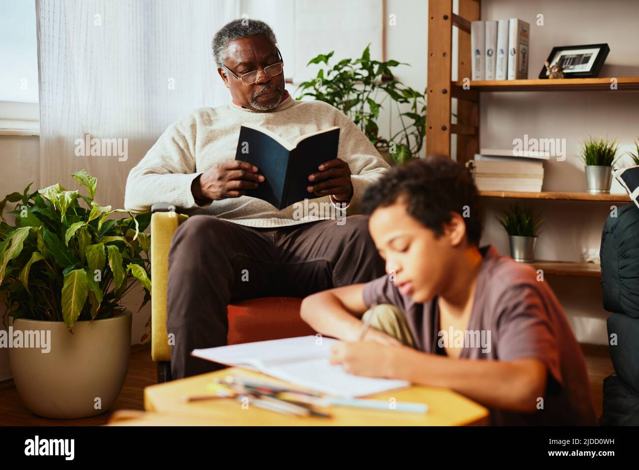 Ein afroamerikanischer Großvater sitzt auf seinem Stuhl und liest ein Buch, während sein geliebter Enkel Hausaufgaben macht. Idyllischer Nachmittag bei den Großeltern. Stockfoto