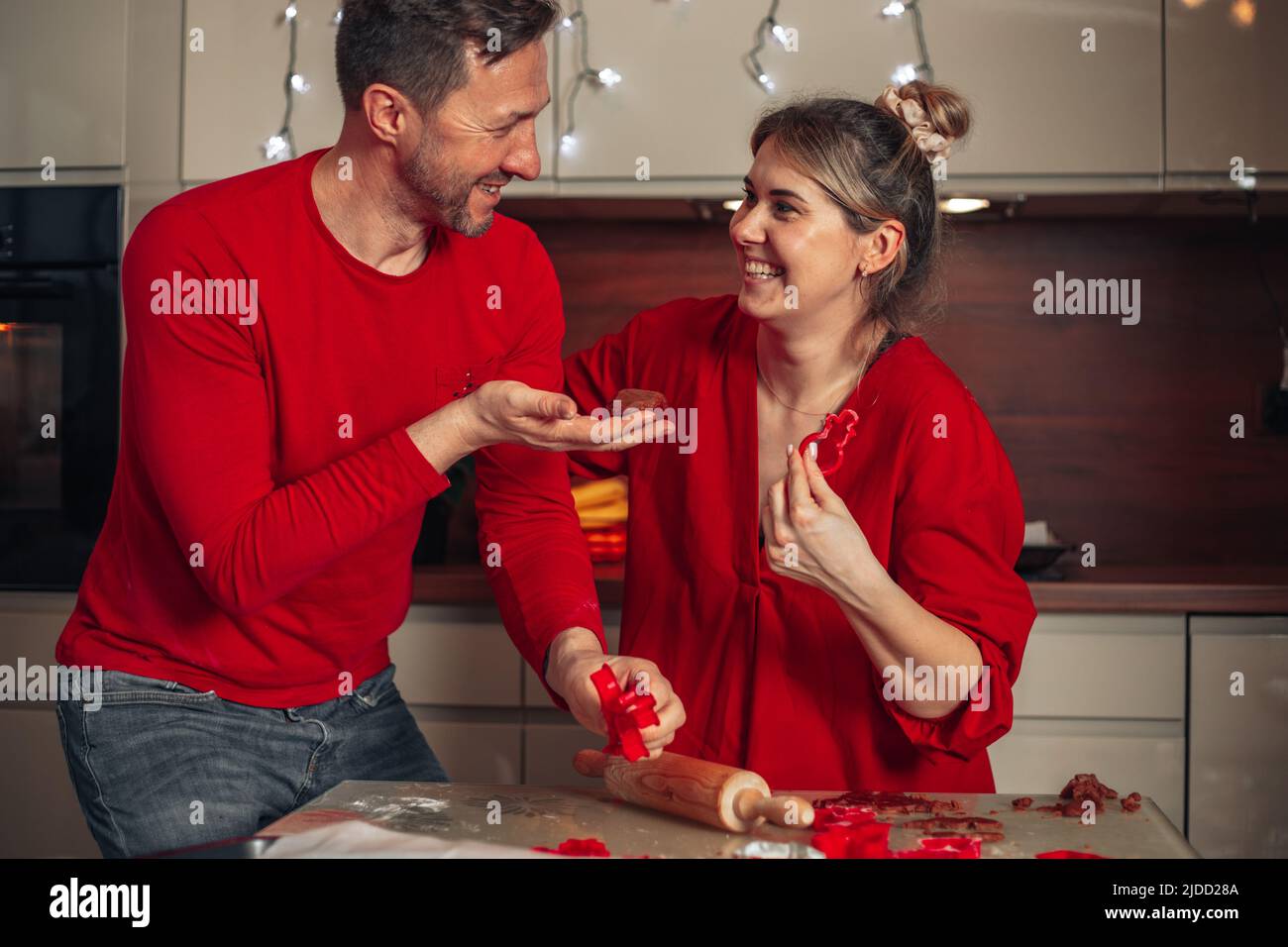 Junge nette Frau und Mann in roten Kleidern lachen fröhlich beim Backen von Keksen. Weihnachtsmomente, glückliches junges Paar. Startseite gemütliche Küche. Stockfoto