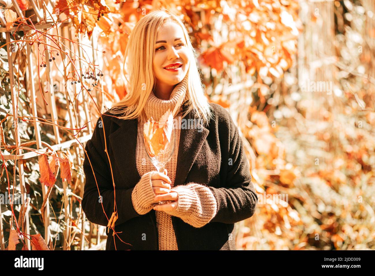 Die schöne blonde 35-Jährige in blackem Kantel und beigefarbenem Pullover hält Glas mit Herbstcocktail in den Händen und genießt einen sonnigen Tag im Stadtpark. Farben Stockfoto