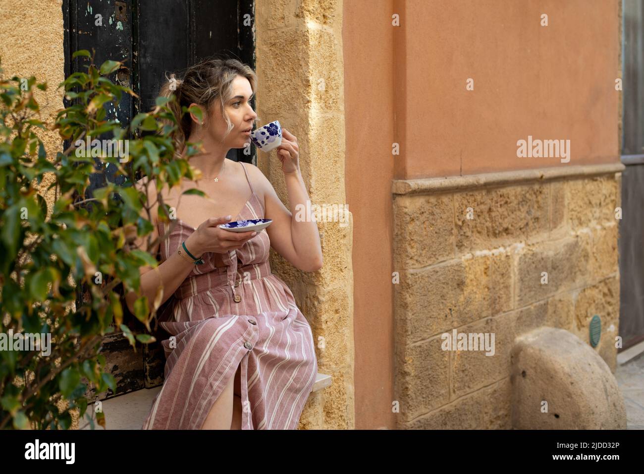 Porträt einer jungen Frau, die im Sommer rosa gestreiftes Kleid trägt, neben dem Olivenbaum sitzt, Kaffee trinkt und das Aroma genießt. Stockfoto