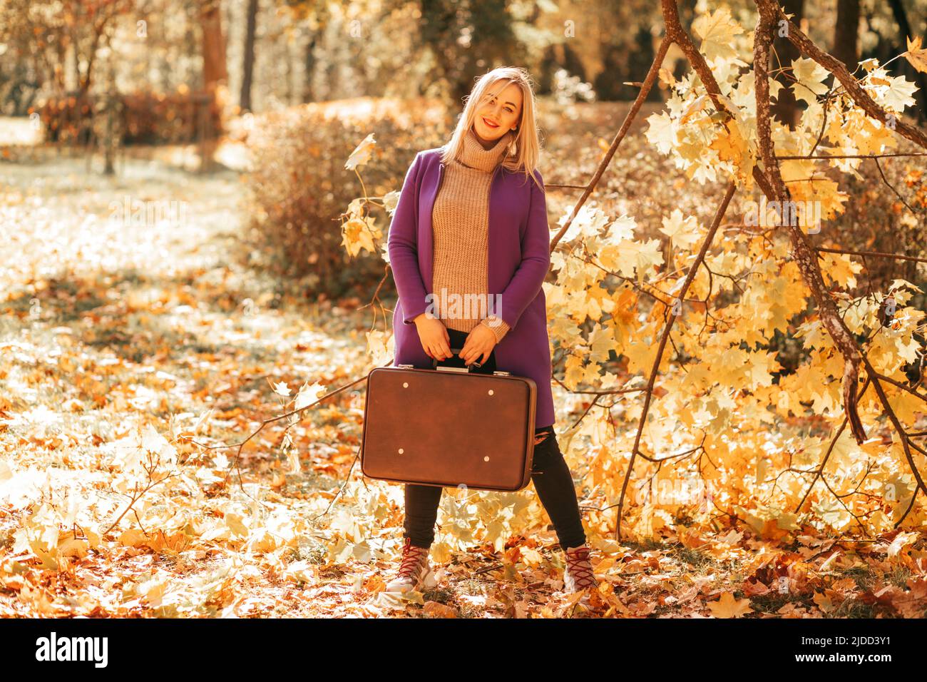 Fröhliche Frau in kuscheliger Kleidung mit altem Koffer in den Händen posiert im Herbstpark voller gefallener Blätter. Herbstfarben. Erholung im Park Stockfoto