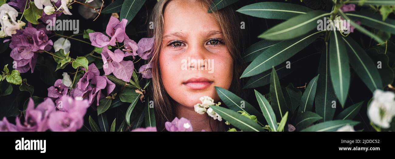 Portrait Gesicht von ehrlichen schönen kleinen Mädchen acht Jahre alt mit braunen Augen auf dem Hintergrund von grünen Pflanzen und rosa Blumen während eines Sommers va Stockfoto