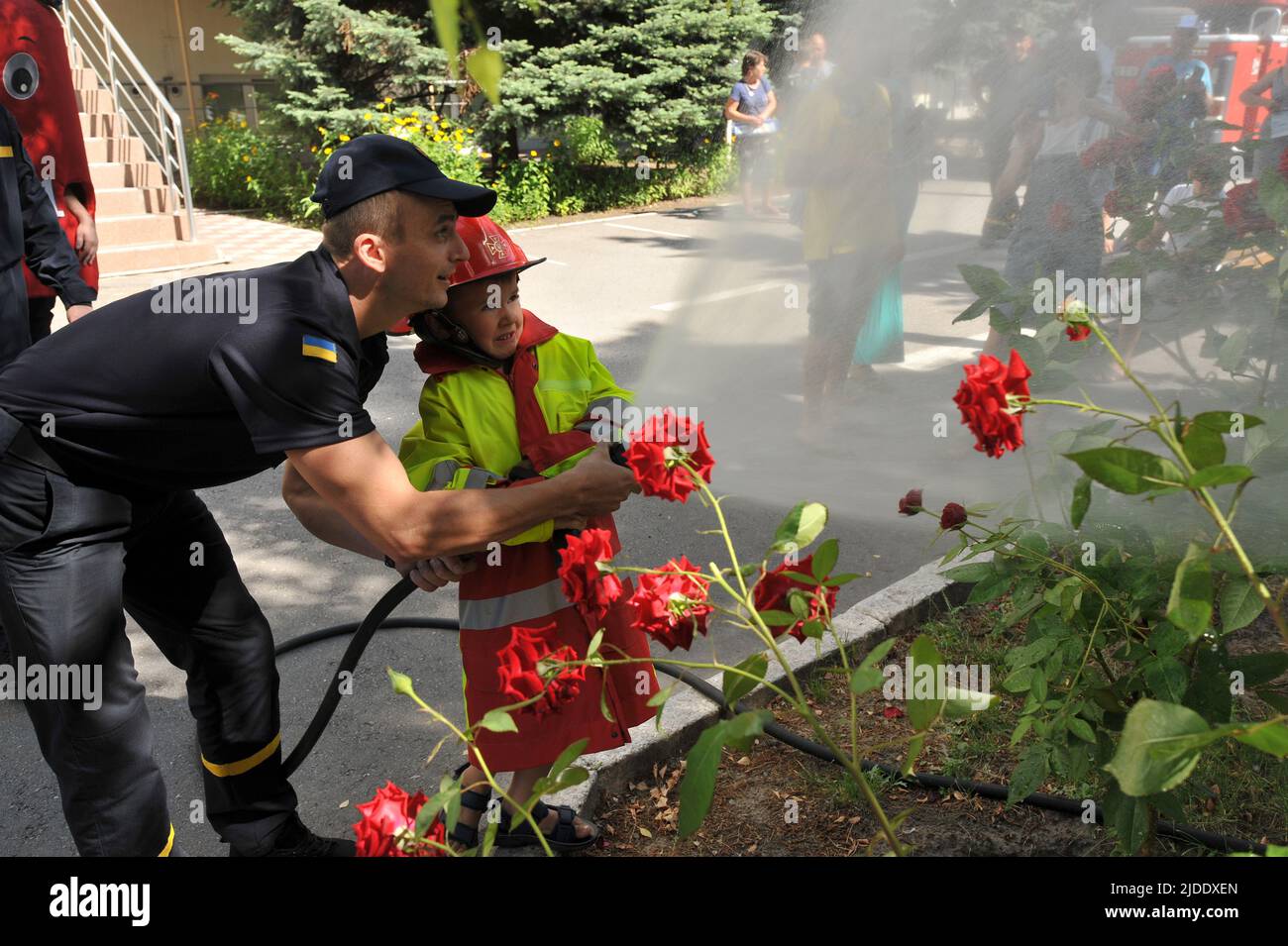 ZAPORIZHZHIA, UKRAINE - 20. JUNI 2022 - Ein Retter hilft einem Jungen, einen Schlauch zu halten und während eines Urlaubs Blumen für intern vertriebene Kinder zu gießen Stockfoto