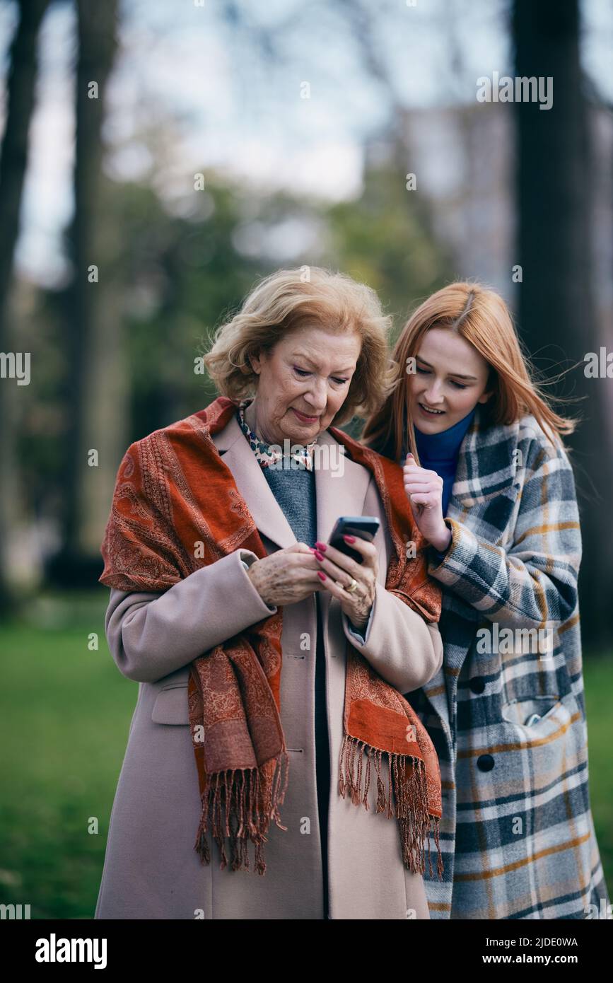 Eine Großmutter stöbert während eines Spaziergangs im Park mit ihrer heranwachsenden Enkelin bei kaltem Wetter durch ihr Telefon. Stockfoto