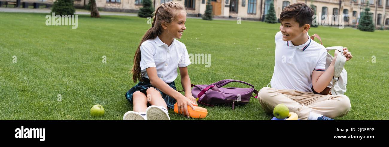 Lächelndes Schulmädchen hält Lunchbox in der Nähe einer asiatischen Freundin mit Rucksack auf Gras im Park, Banner Stockfoto