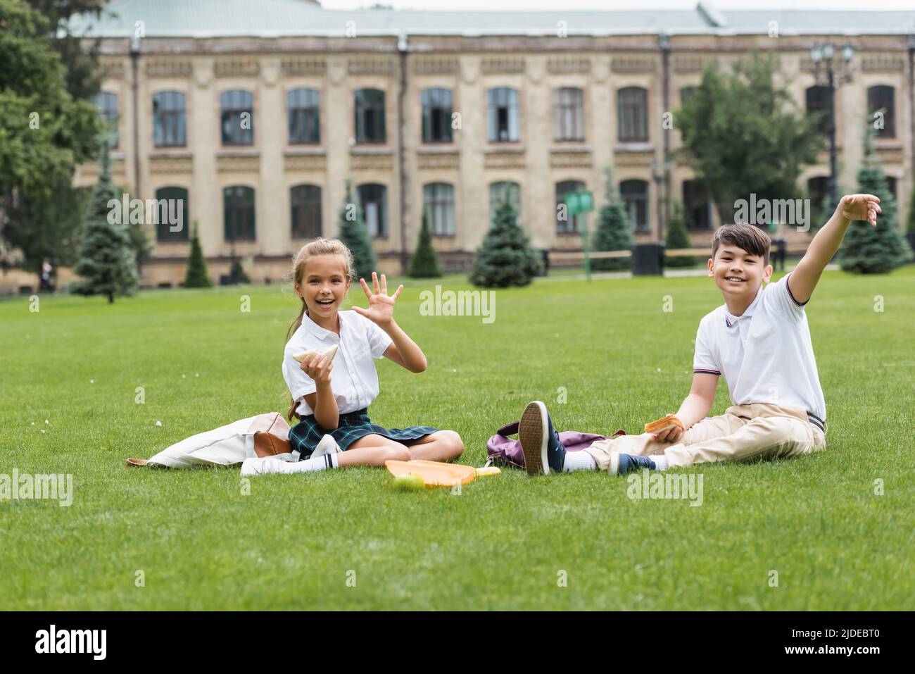 Positive Schülerin hält Sandwich und winkt Hand an der Kamera in der Nähe von asiatischen Freund auf Rasen im Park Stockfoto