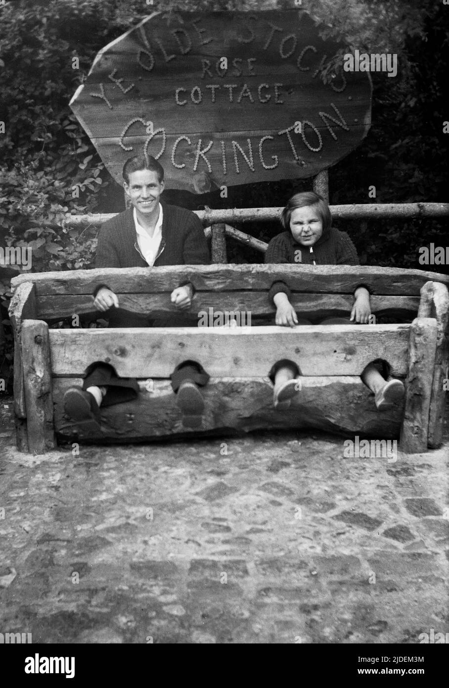 1935, historisch, ein Vater und eine Tochter, die für ihr Foto in „Ye Olde Stocks“ im Rose Cottage in Cockington, Torquay, Devon, England, Großbritannien, sitzen. Vorräte waren Holzgeräte, die als eine Form der "Bestrafung" und "Scham" für lokale Straftäter von Kleinkriminalität verwendet wurden und die Straftäter mit Füßen und Händen fesselten. Auf Dorfgrüns in Großbritannien gesehen, waren sie bis zum Ende des 18.. Jahrhunderts im Einsatz. Stockfoto