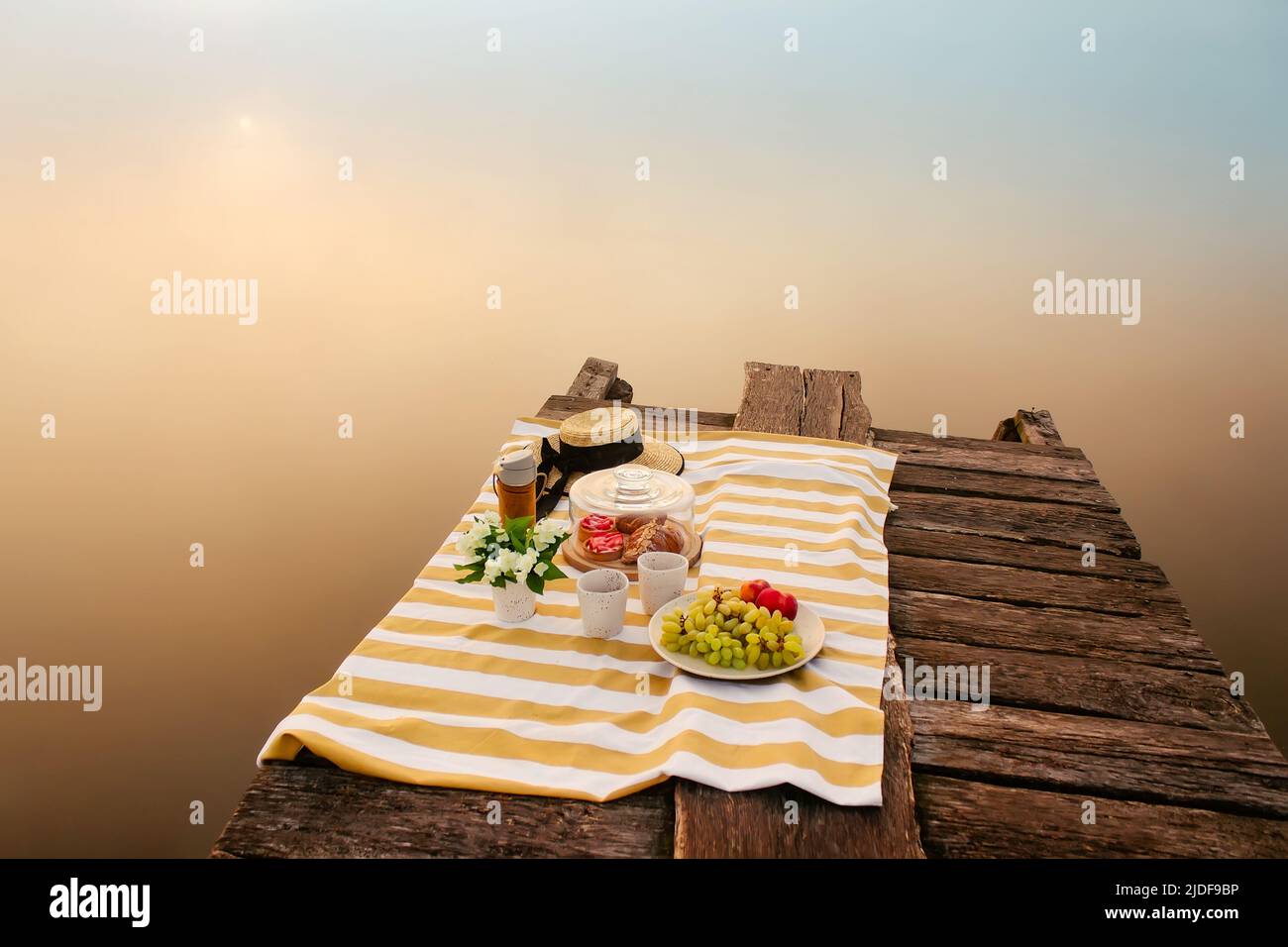 Picknick mit leckerem Essen und Kaffee auf einer Holzbrücke in der Nähe des wunderschönen nebligen Sees bei Sonnenaufgang Stockfoto
