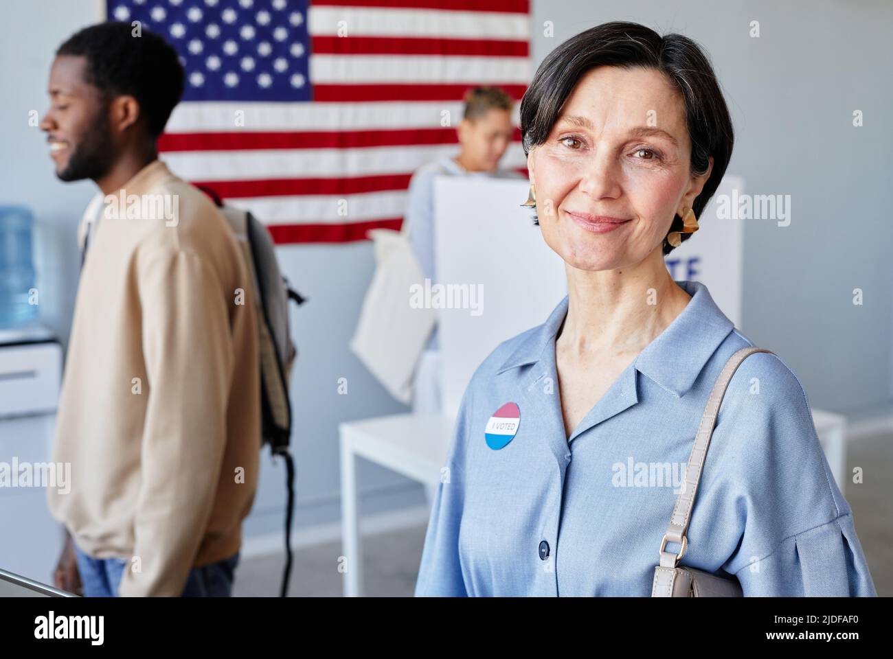 Porträt einer lächelnden erwachsenen Frau an der Wahlstation, die mit der amerikanischen Flagge im Hintergrund und dem Kopierbereich lächelt Stockfoto