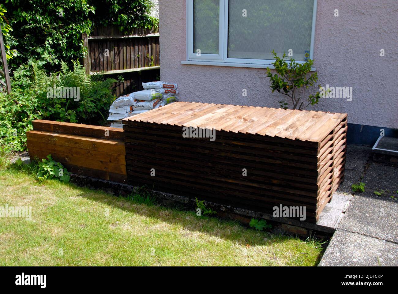 Stapel von hölzernen Zaunplatten, Pfosten, Kiesbretter und Zementmischung, auf der Terrasse gelagert, bereit für den Ersatz des alten häuslichen Gartenzauens Stockfoto