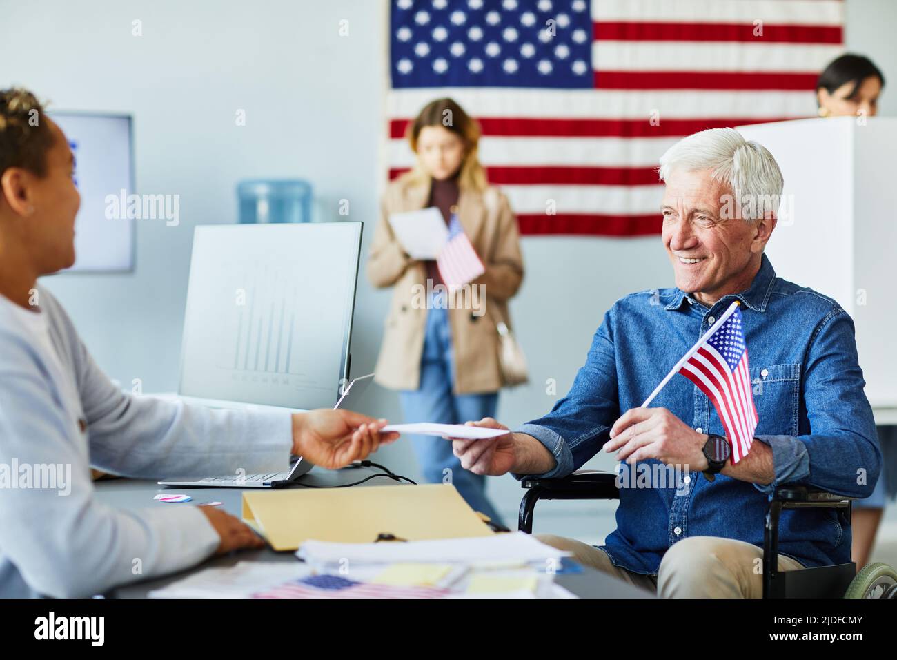 Porträt eines lächelnden, behinderten älteren Mannes, der am Wahltag in der Wahlstation den Stimmzettel mit US-Flagge im Hintergrund erhielt Stockfoto