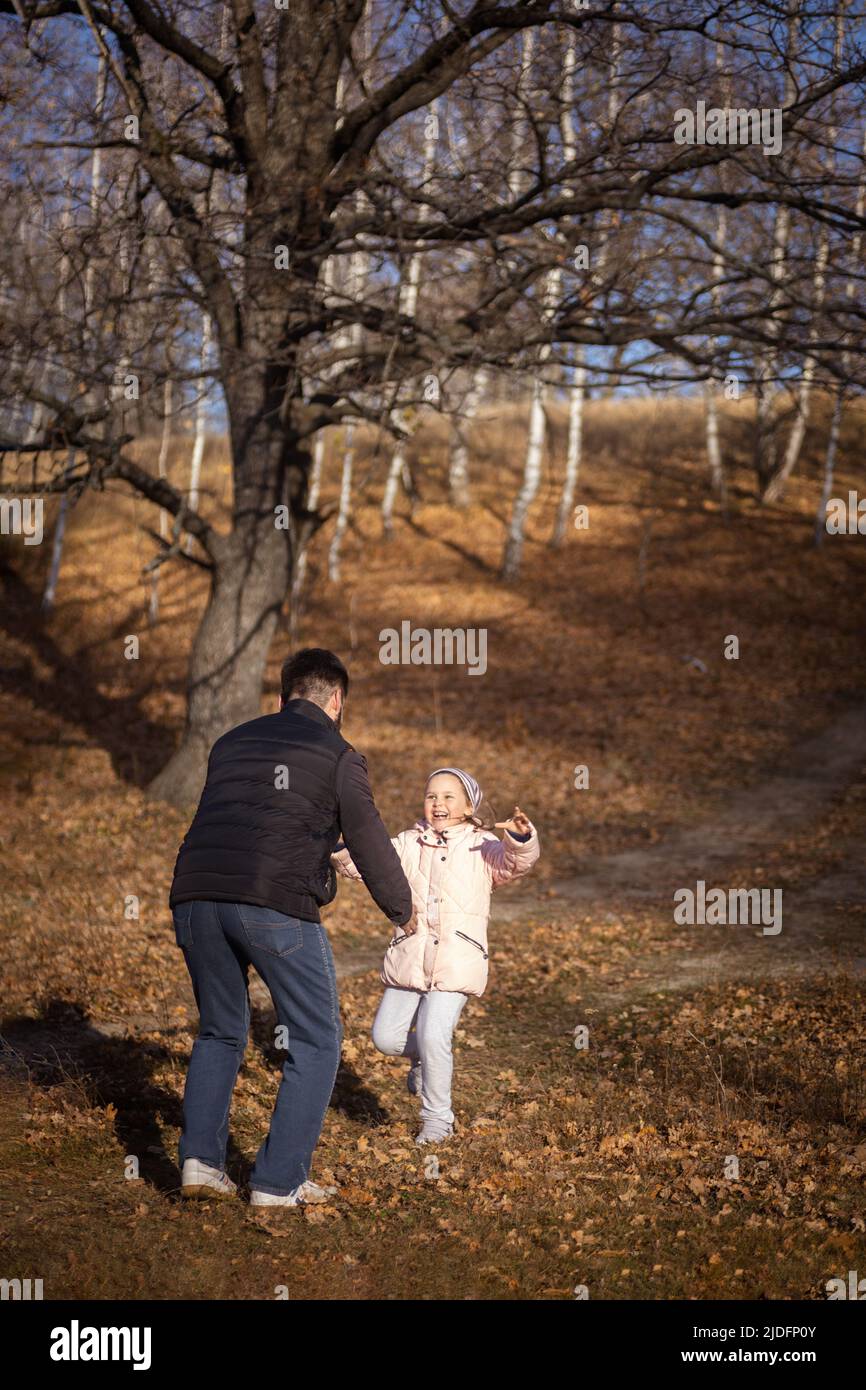Liebevoller Vater, der mit der kleinen Tochter spielt, Spaß hat und lächelt, wenn er am sonnigen Herbsttag im Wald mit trocken gefallenen Blättern auf dem Boden spazieren geht Stockfoto