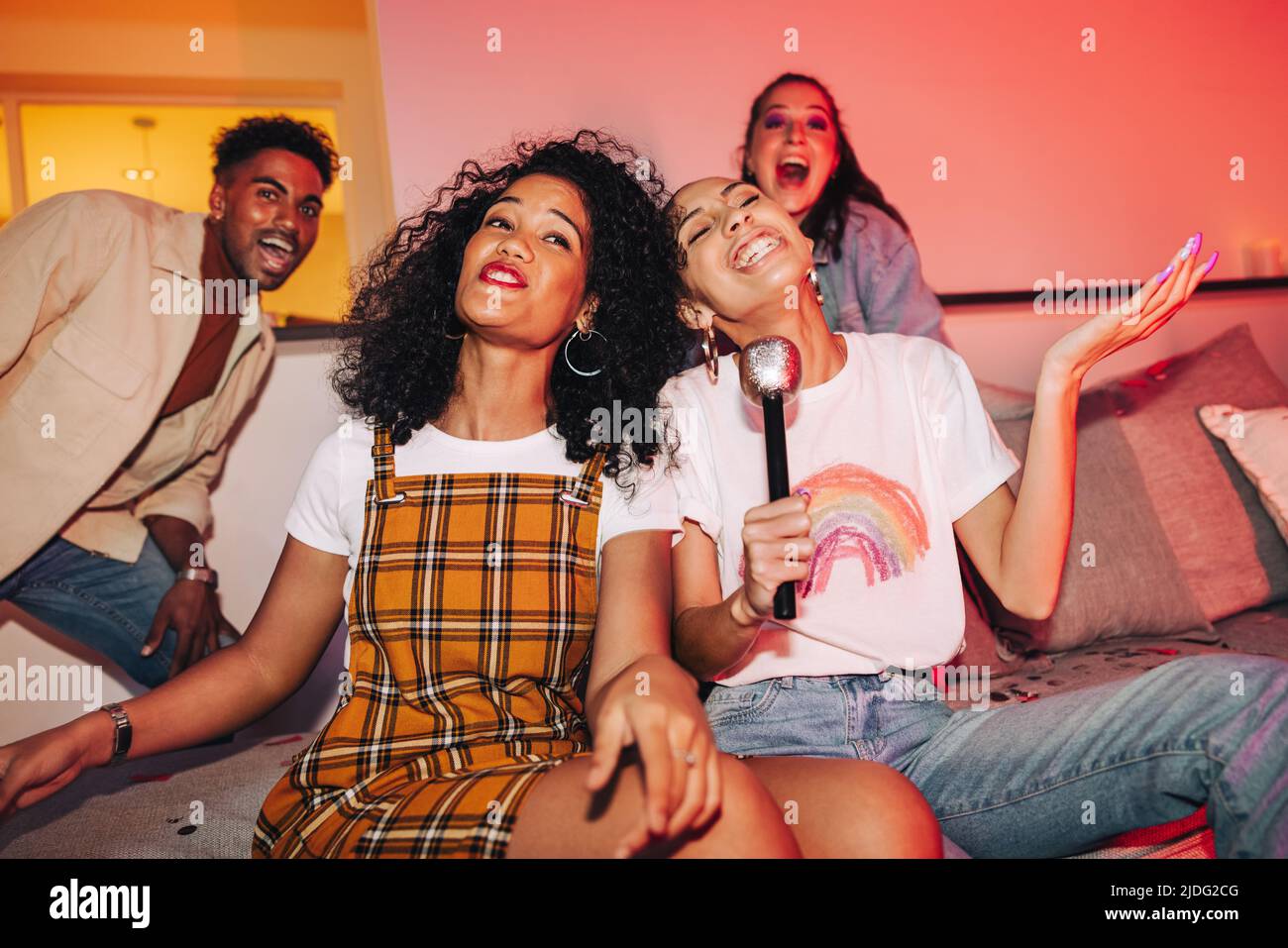 Zwei junge Frauen unterhalten ihre Freunde am Karaoke-Abend. Fröhliche Freundinnen singen ihr Lieblingslied auf einer Hausparty. Gruppe der Multikulturalität Stockfoto