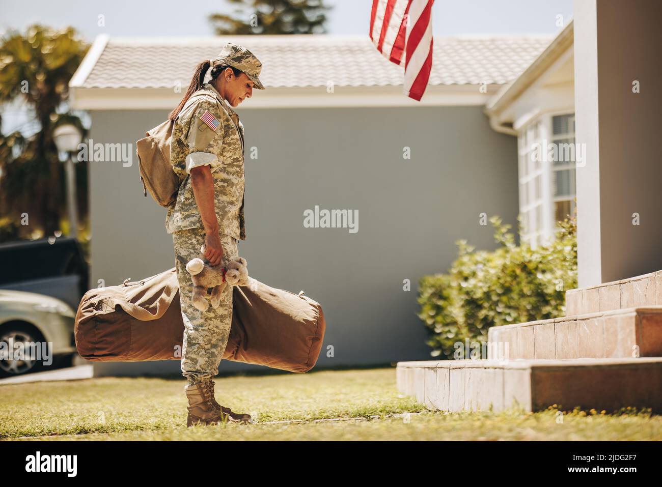 Patriotische Militärmutter, die einen Teddybär in der Hand hält, während sie mit ihrem Gepäck vor ihrem Haus steht. Amerikanische Soldatin, die nach dem Dienst h nach Hause zurückkehrt Stockfoto