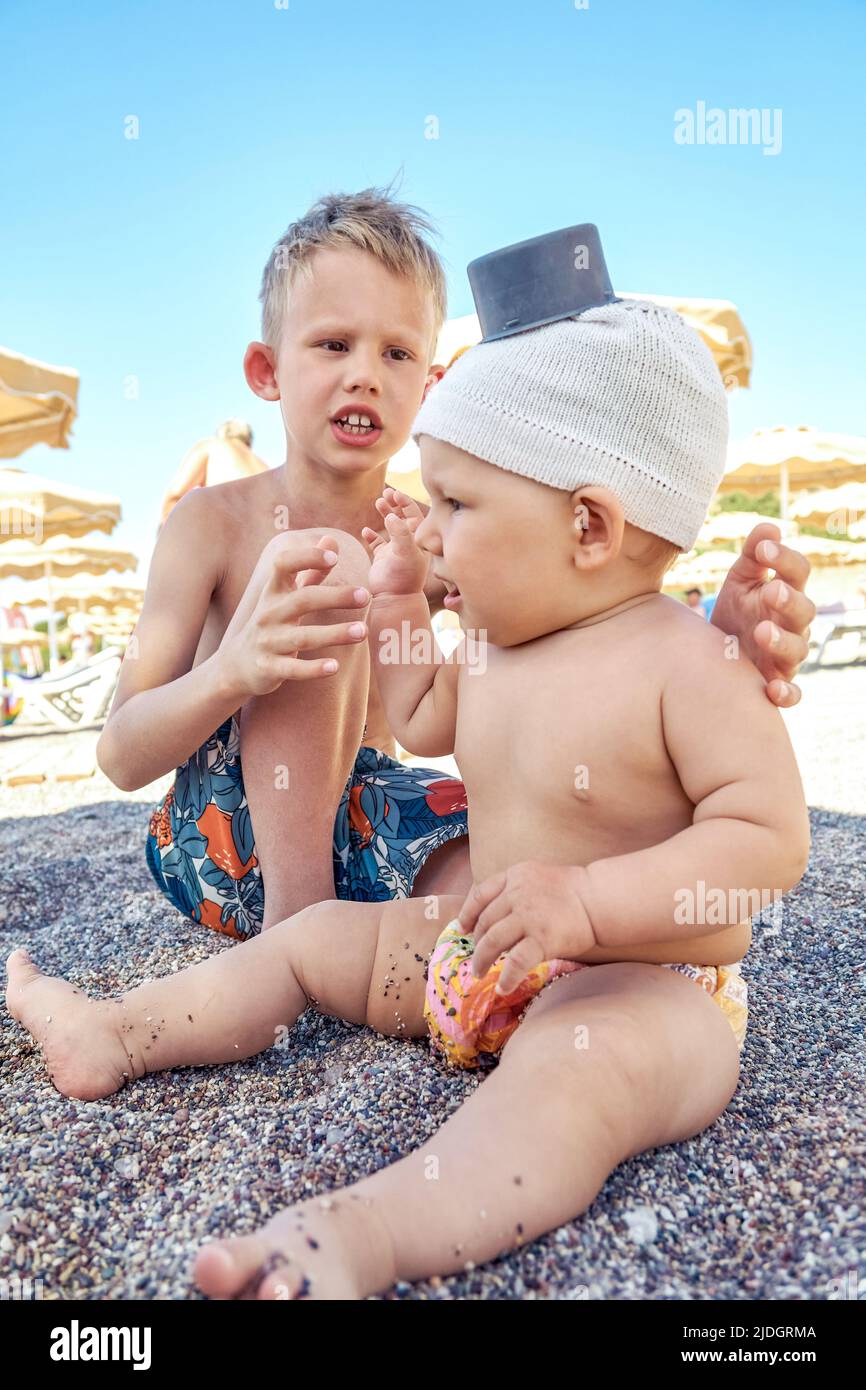 Der ältere Bruder spielt mit dem Geschwisterchen, das auf Sand im Schatten des Regenschirms sitzt. Blonde Schülerin und Kleinkind im Hut genießen Urlaub am Strand Stockfoto
