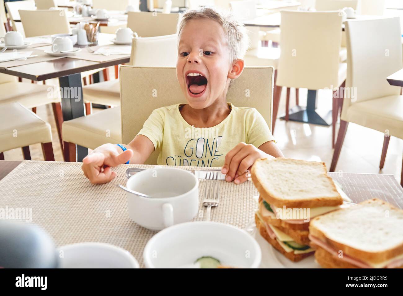 Der blonde Vorschuljunge lacht beim Frühstück im Hotel. Netter Junge hat Spaß am Tisch zu sitzen und große Sandwiches im Sommerurlaub zu essen Stockfoto