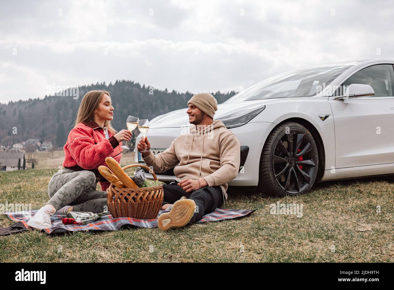 Lächelndes junges, modernes Paar, das eine Reise in die Natur macht, Wein und Trauben genießt, während es entspannt neben ihrem neuen exklusiven Elektroauto sitzt Stockfoto