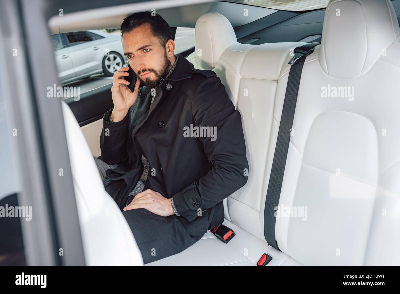 Kaukasischer junger erfolgreicher Mann, der ernsthaft auf dem Smartphone spricht, während er auf dem Rücksitz eines fahrenden Autos sitzt Stockfoto