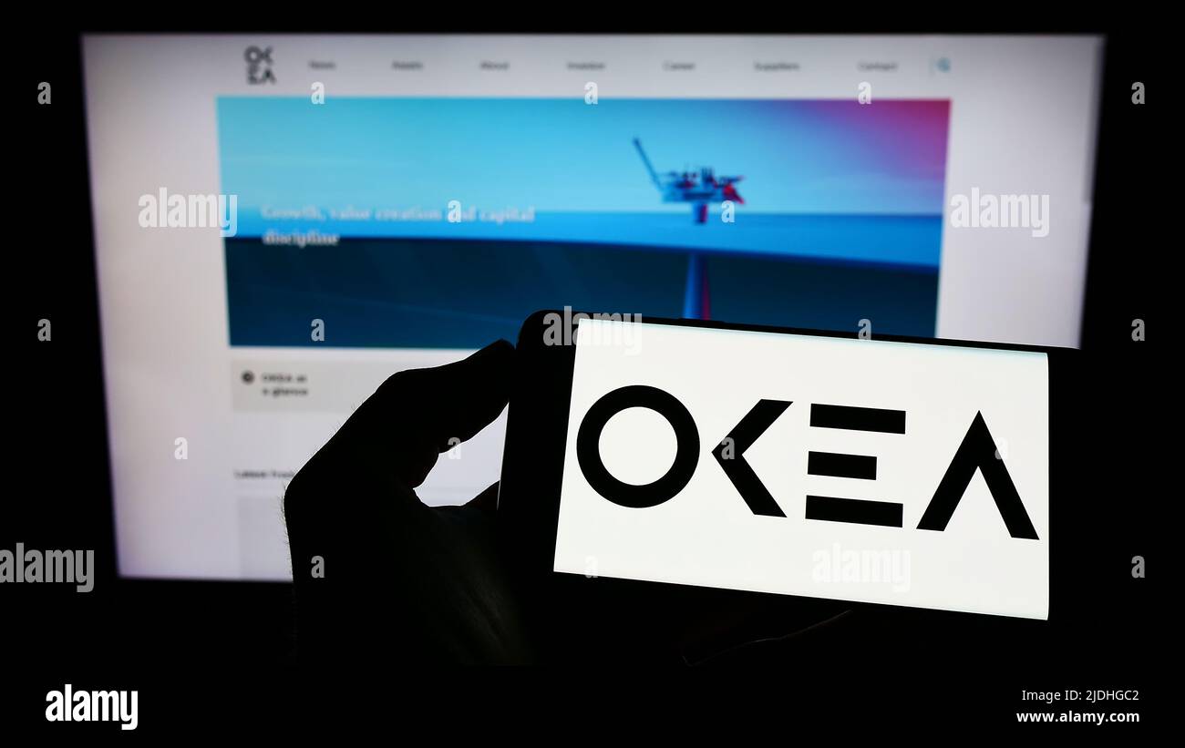 Person, die Mobiltelefon mit dem Logo des norwegischen Ölunternehmens OKEA ASA auf dem Bildschirm vor der Business-Webseite hält. Konzentrieren Sie sich auf die Telefonanzeige. Stockfoto