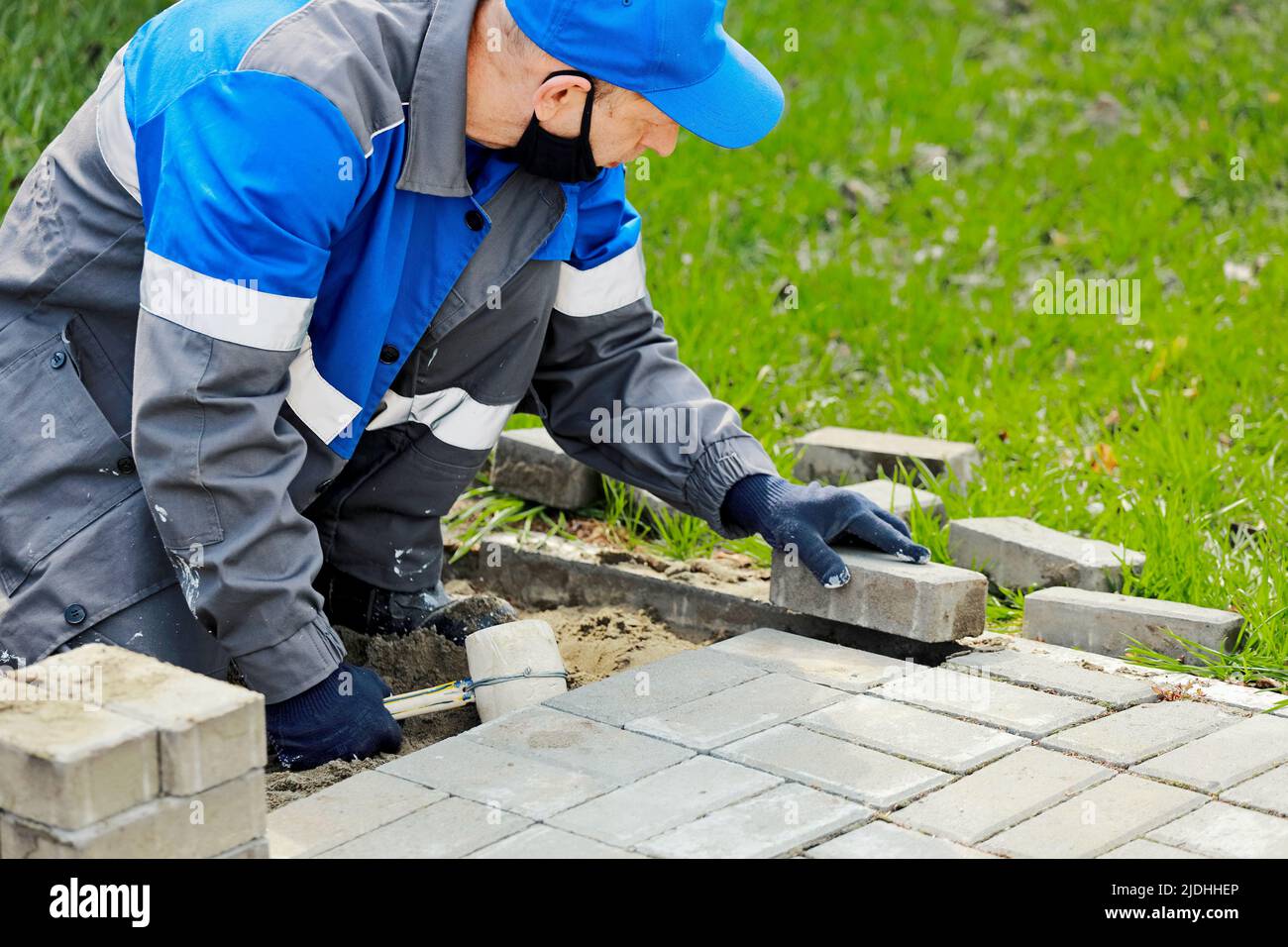 Maurer in Arbeitskleidung sitzt auf dem Bürgersteig und legt Pflasterplatten aus. Anblick eines arbeitenden Mannes im Freien. Der professionelle Baumeister macht die Anordnung des Territoriums Sommertag. Echte Szene. Stockfoto