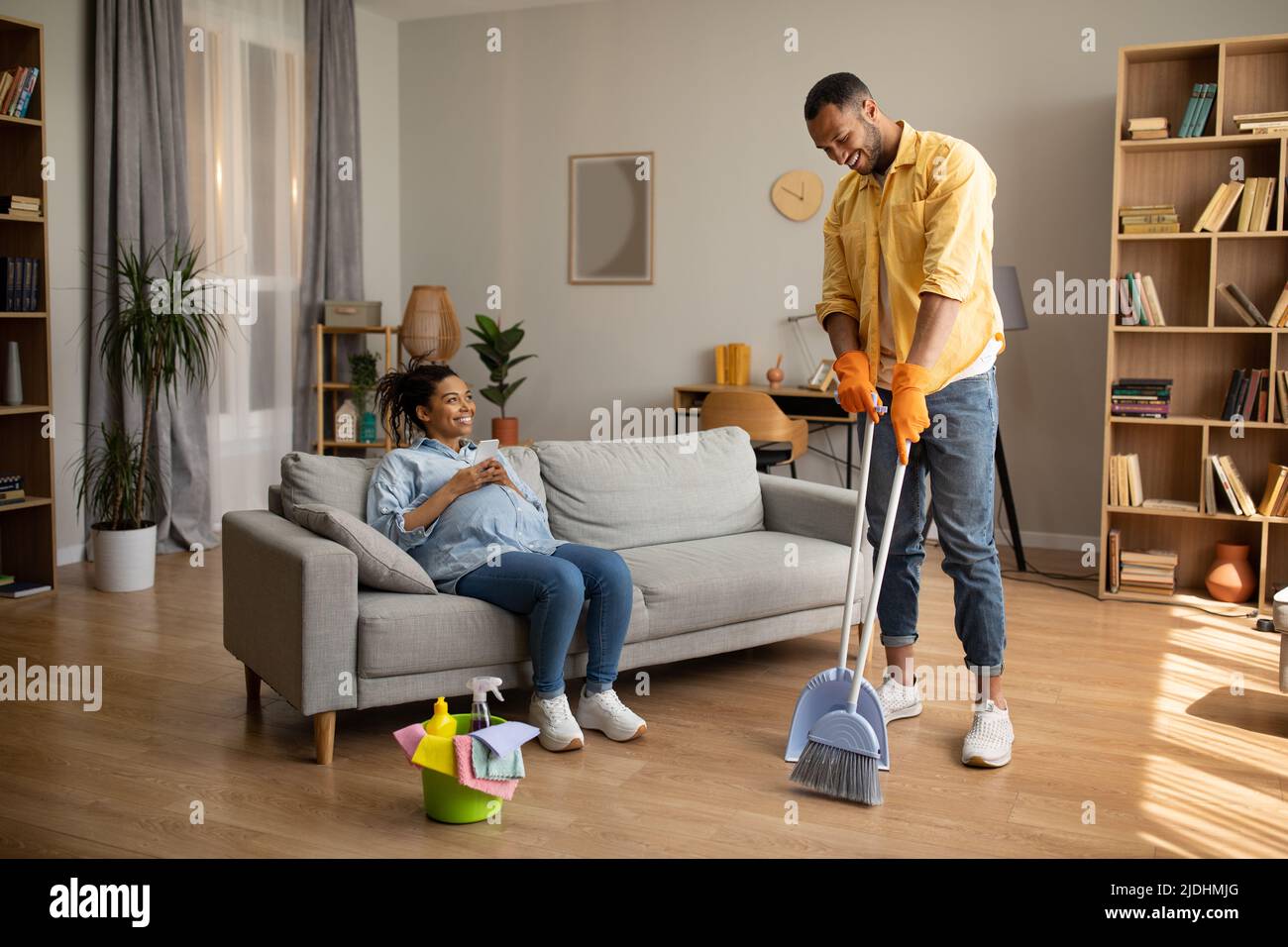 Afrikanischer Ehemann, Der Haus Putzt, Während Seine Frau Zu Hause Das Mobiltelefon Benutzt Stockfoto