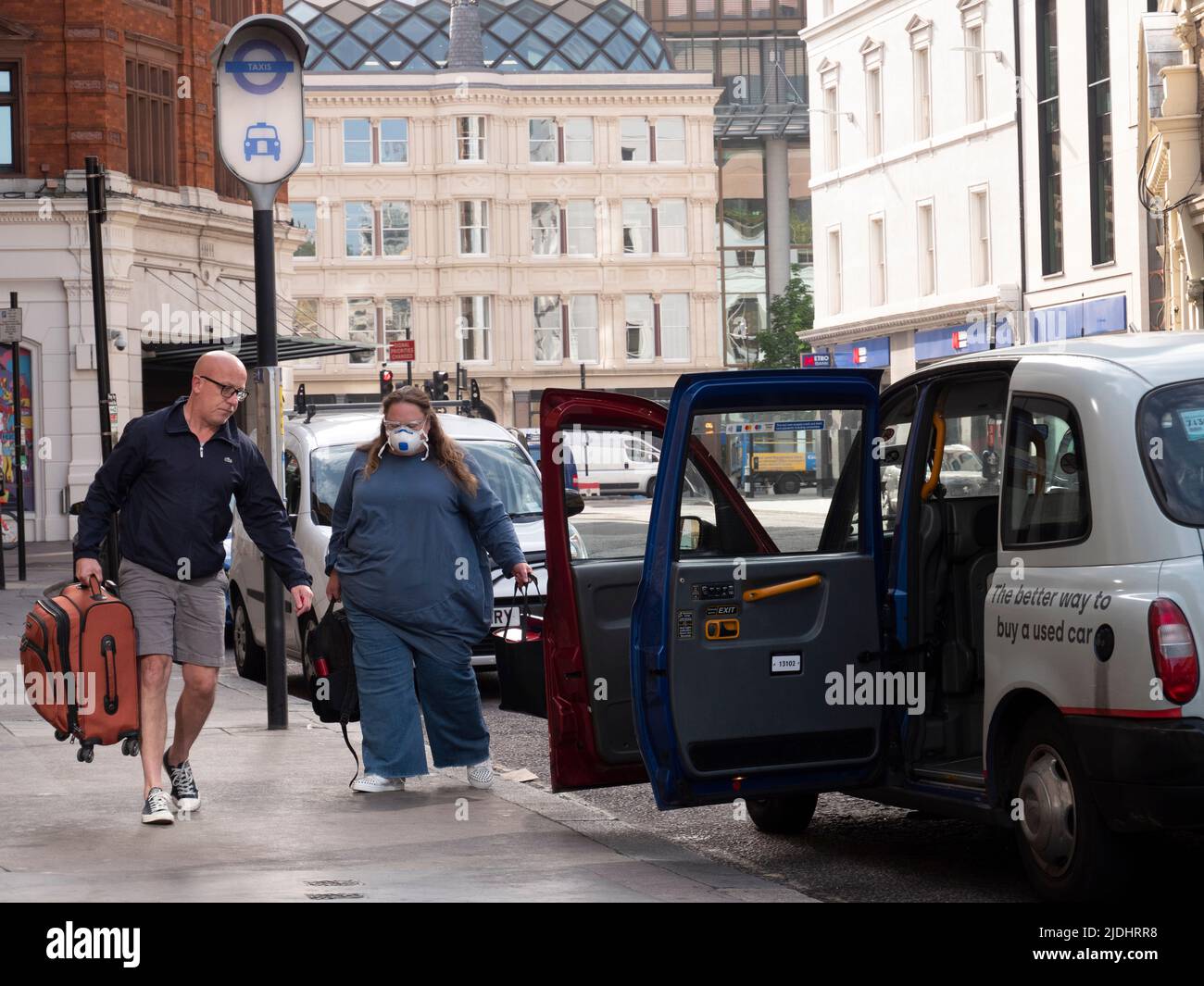 Die schwarze Taxifahrerin in London hilft, Gepäck in das wartende Taxi zu laden, wobei die Frau eine PSA-Covid-Maske trägt Stockfoto