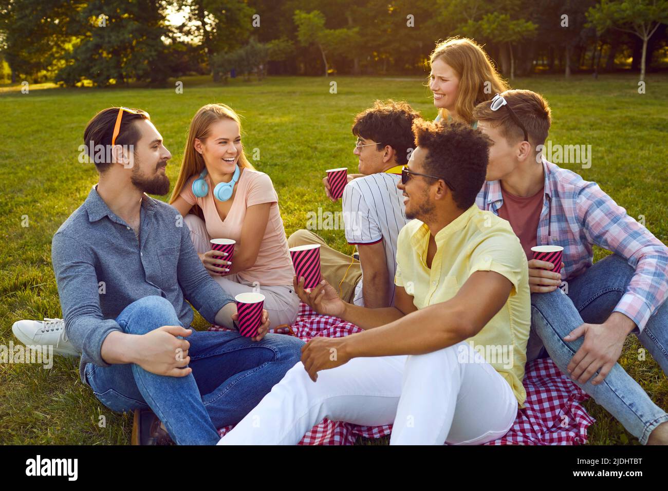 Fröhliche multiethnische Freunde sitzen am sonnigen Sommertag auf grünem Rasen mit Pappbechern. Stockfoto