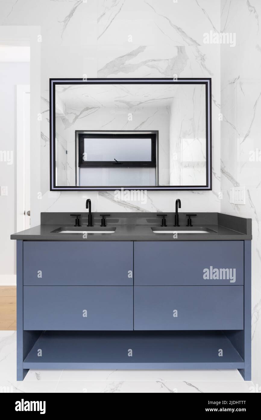 Ein luxuriöses Badezimmer mit mit mit Marmorfliesen bedeckten Wänden, blauem Schrank, dunkelgrauem Schminktisch und einem Spiegel mit Licht am Rahmen. Stockfoto