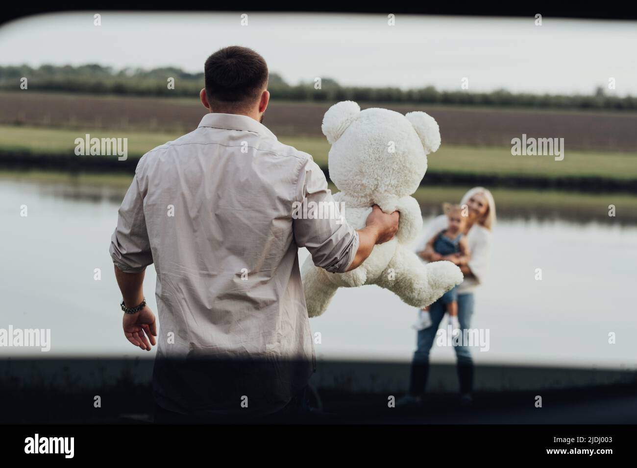 Der junge Vater hielt den Teddybären in der Hand vor seiner Familie, und Vater bereitete ein Geschenk für seine kleine Tochter, Blick durch das Autofenster, vor Stockfoto