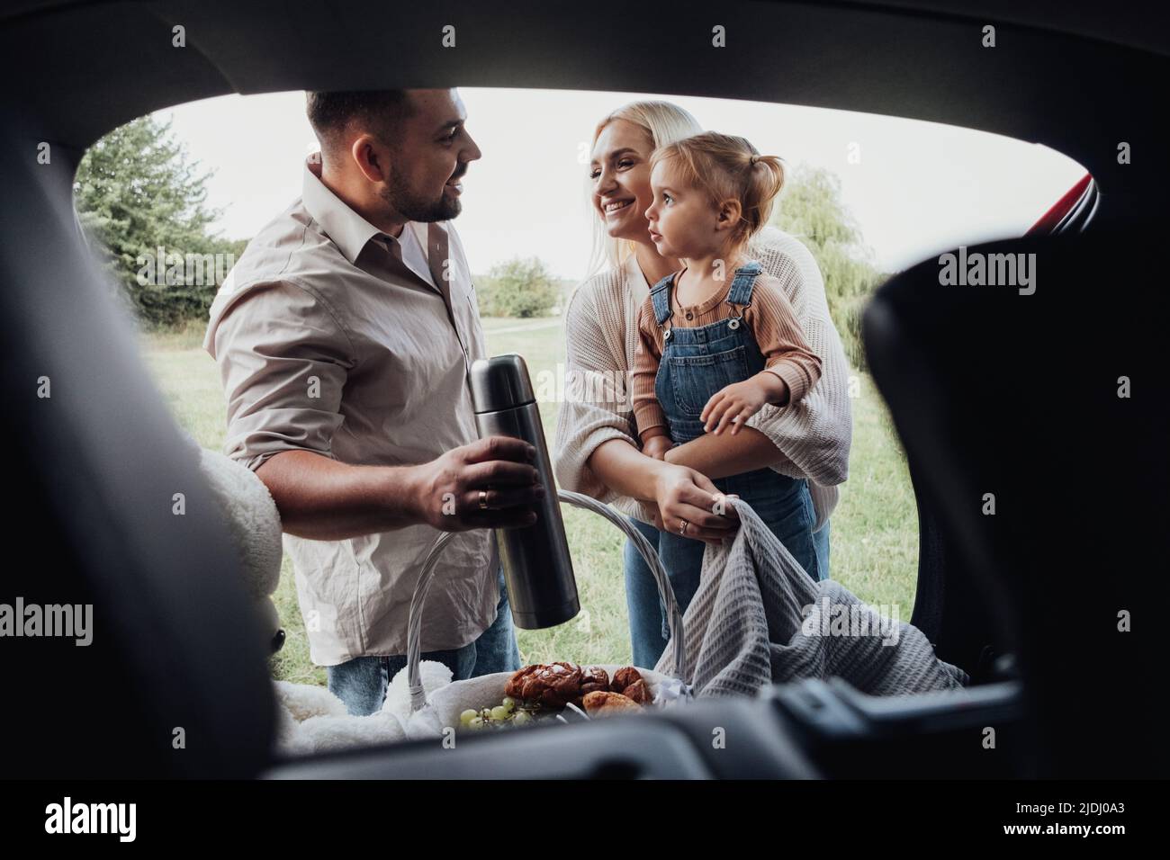 Junge Familie, zwei Eltern mit ihrem kleinen Kind, die sich auf ein Picknick im Freien vorbereiten, Blick durch den offenen Kofferraum des SUV-Autos Stockfoto