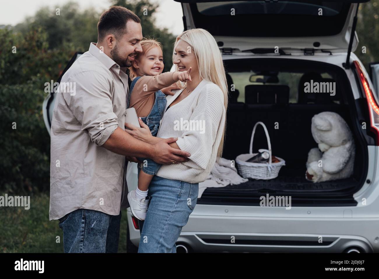 Junge Eltern mit ihrer Kleinkindtochter umarmen sich vor dem SUV-Auto, Familie genießt Roadtrip-Wochenende Stockfoto