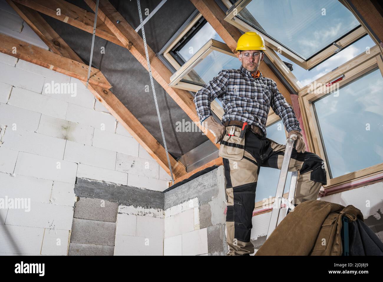 Installation von Dachfenstern in einem neu gebauten Wohnhaus. Beschäftigter kaukasischer Baumeister bei seinem 40s auf einer Leiter im Gebäude der Mitte eines stehend Stockfoto