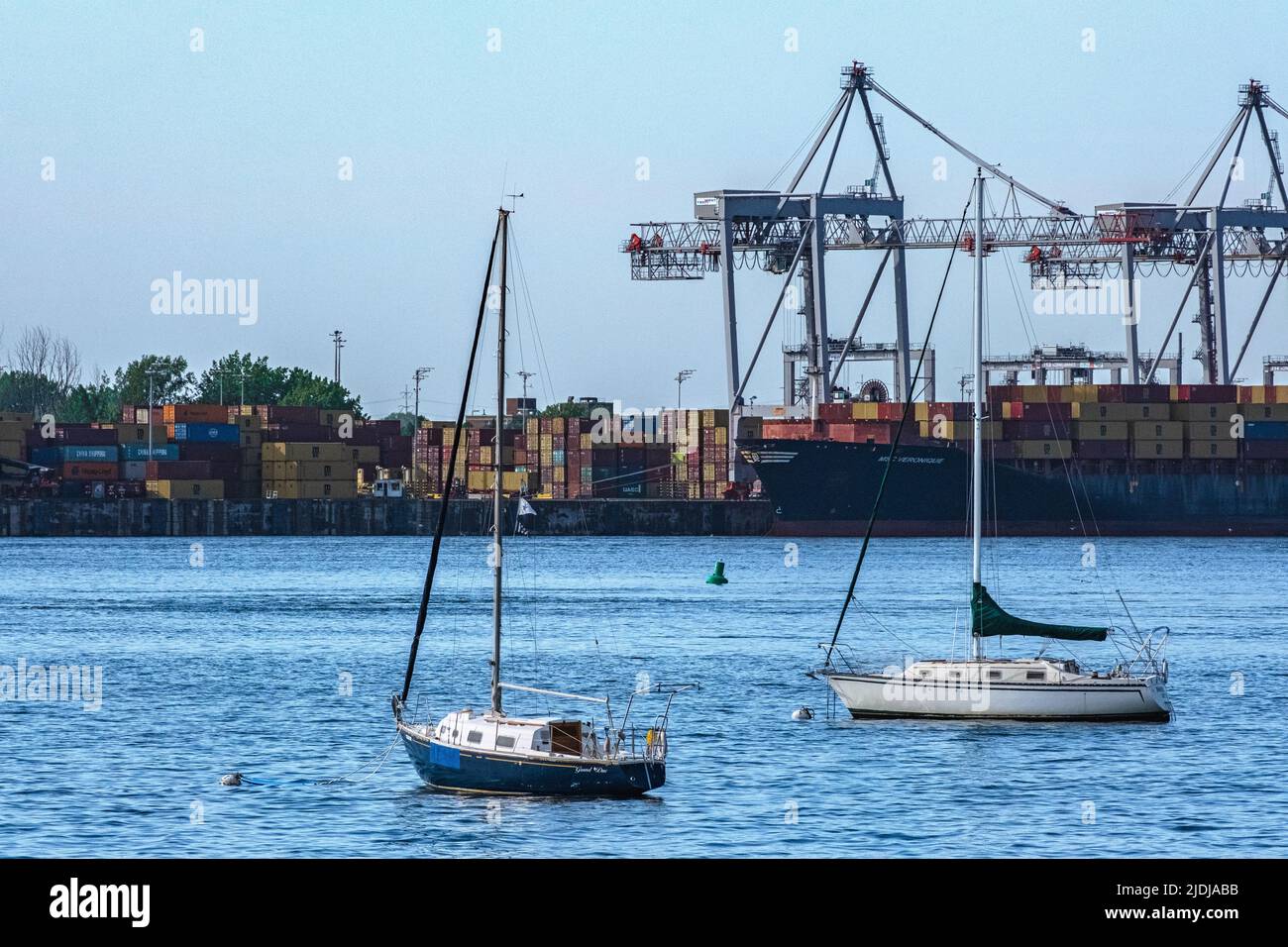 Zwei Segelboote fahren am heißen Tag am Hafen von Montreal ein. In der Ferne werden die Transportbehälter von Kränen gestapelt, wenn ein Schiff vorbeifährt. Stockfoto