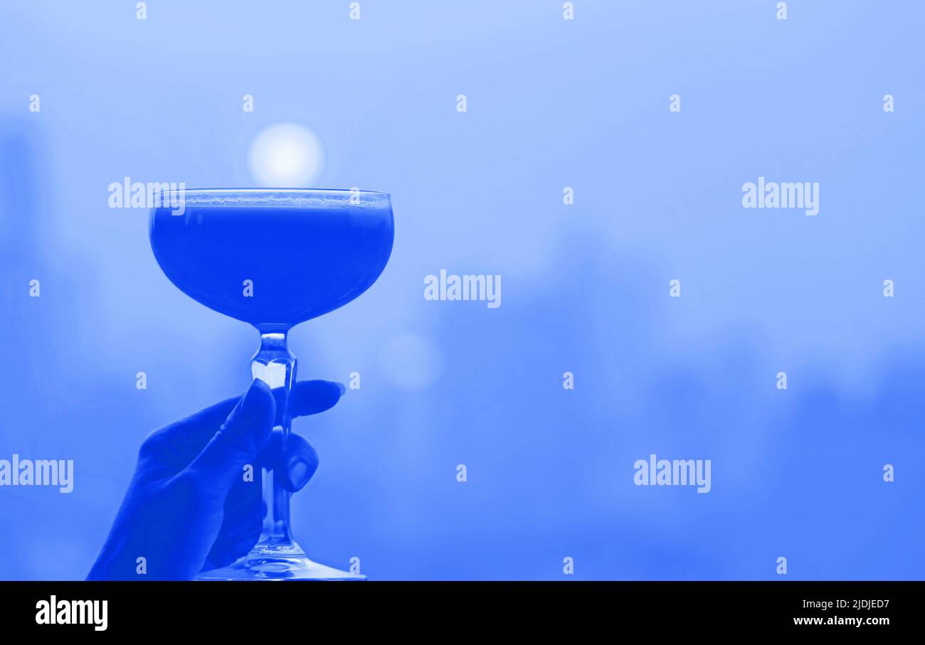 Kobaltblaues Cocktailglas im Pop-Art-Stil in der Hand mit verschwommenem Blick auf die Stadt im Hintergrund Stockfoto