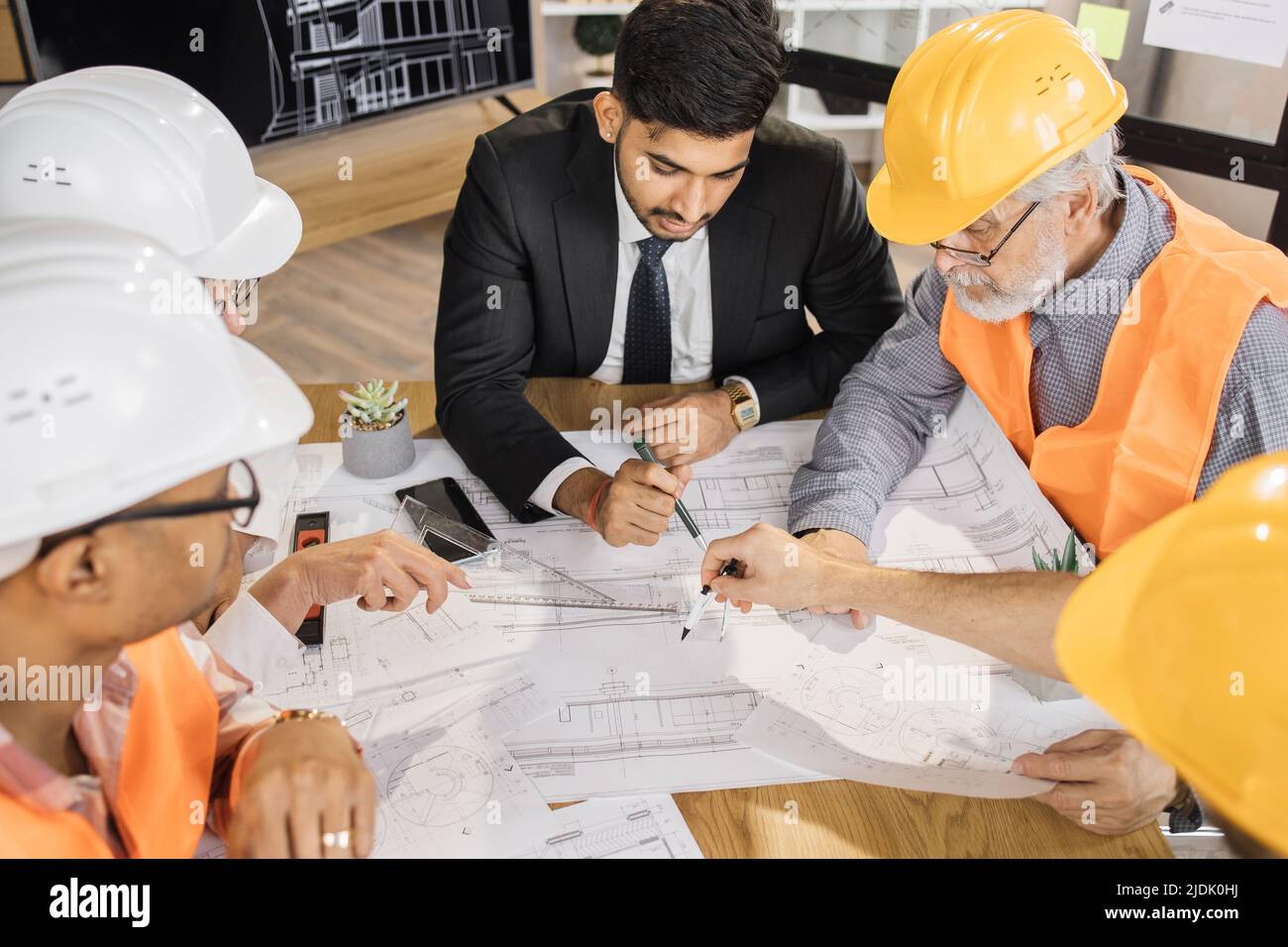 Qualifizierte Architekten, Ingenieure und Designer mit Geschäftstreffen im Büro. Multikulturelle Kollegen in Anzügen und Helmen sitzen am Tisch und schauen sich Blaupausen an. Konstruktionskonzept. Stockfoto