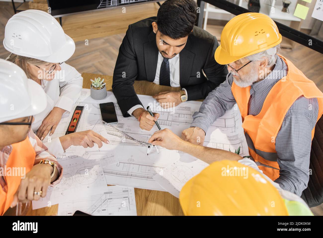 Qualifizierte Architekten, Ingenieure und Designer mit Geschäftstreffen im Büro. Multikulturelle Kollegen in Anzügen und Helmen sitzen am Tisch und schauen sich Blaupausen an. Konstruktionskonzept. Stockfoto